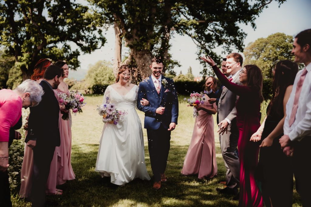 Bride and groom smiling as they walk arm in arm through a tunnel of cheering guests throwing confetti, surrounded by bridesmaids and friends on a sunny day at Barns and Yard.